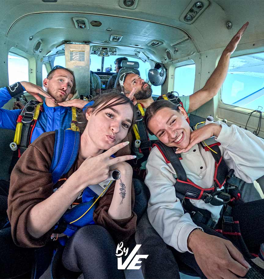 Groupe de personnes assises dans un avion, équipées pour un saut en parachute, posant de manière amusante pour la caméra.