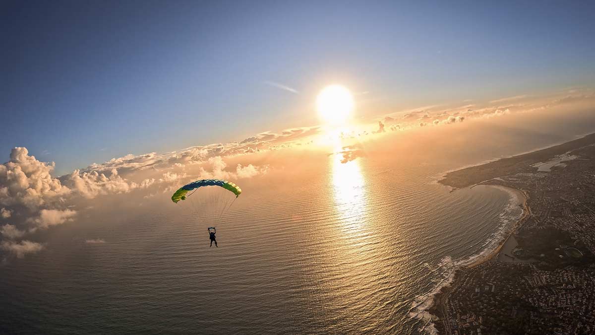 Un parachutiste descend sous voile au‑dessus de la mer, face au soleil couchant, avec la côte visible sur la droite.