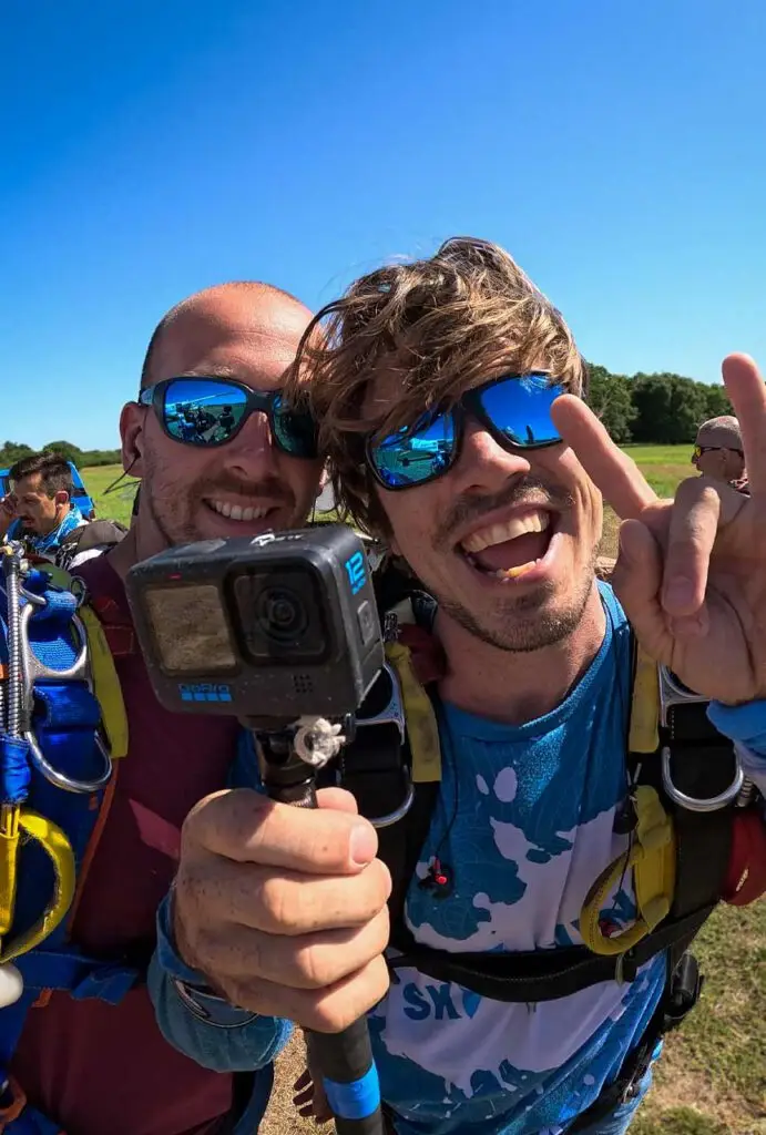 Deux personnes en tenue de parachutisme posant avec une caméra, souriantes et portant des lunettes de soleil réfléchissantes.