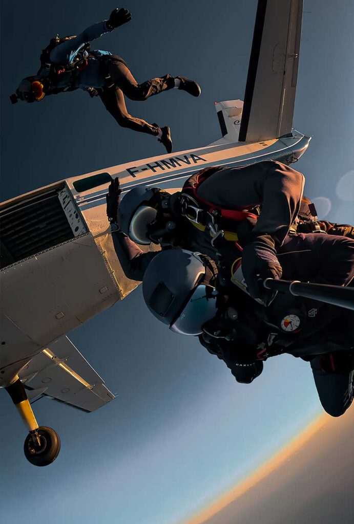 Duo en saut tandem quittant un avion en vol, avec un caméraman qui s’élance derrière eux, dans une lumière de fin de journée.