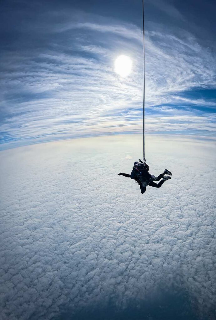 Duo en chute libre au‑dessus d’une couche nuageuse dense, avec le soleil perçant un ciel strié de nuages.