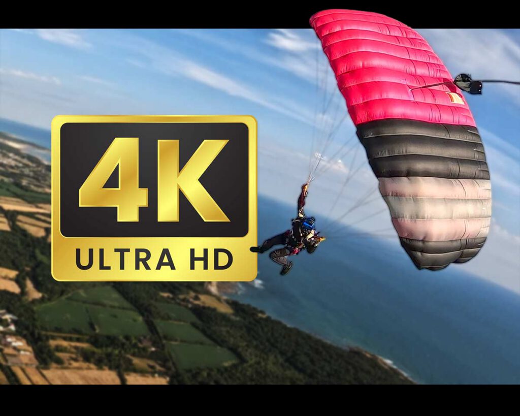 Parachutiste sous une voile rose et noire en descente au‑dessus du littoral, avec un logo « 4K Ultra HD » au premier plan.