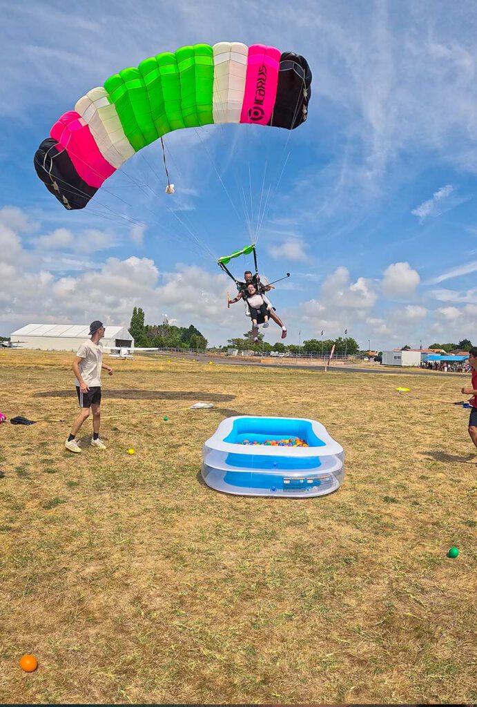 Duo en parachute sous une voile multicolore se posant près d’une petite piscine gonflable remplie de balles, avec des personnes au sol.