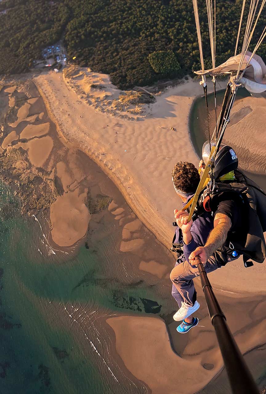 Duo en parachute sous voile survolant une plage, des dunes et des bancs de sable avec une eau peu profonde.